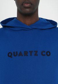 Felpa blu con cappuccio caratterizzata da un logo ricamato sul fronte "QUARTZ CO" in nero. Tessuto morbido con una vestibilità comoda e cappuccio con lacci.