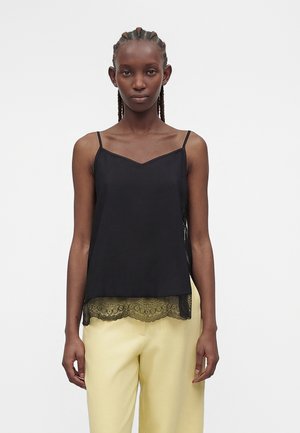 Kvinde med fletninger iført en sort camisole med blonderkant og blege gule bukser, der står mod en ensfarvet hvid baggrund.