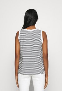 Personne aux longs cheveux noirs portant un haut sans manches à rayures noir et blanc et un pantalon blanc, vue de dos sur un fond uni.