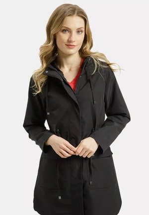 Blondynka z falistymi włosami w czarnej kurtce z kapturem, założonej na czerwony top, stoi z rękami splecionymi w talii na białym tle.