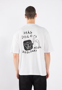 White cotton t-shirt featuring a black graphic on the back with the text “Más disco, menos problemas” and illustrations of a butterfly and earbuds.