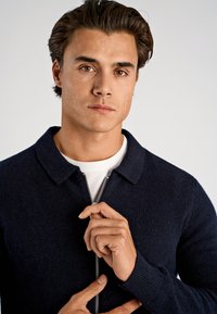 Granatowy sweter zapinany na zamek z kołnierzem, wykonany z fakturowanego dzianinowego materiału. Model trzyma zamek, pod spodem ma na sobie białą koszulę.