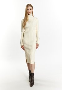 Ribbed, cream-colored knit dress with a high collar, side slits, and buttoned cuff details. Worn with dark brown ankle boots.