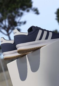 Zapatillas Adidas azul marino con rayas blancas y suelas de goma descansando sobre un pedestal blanco al aire libre con árboles desenfocados al fondo.