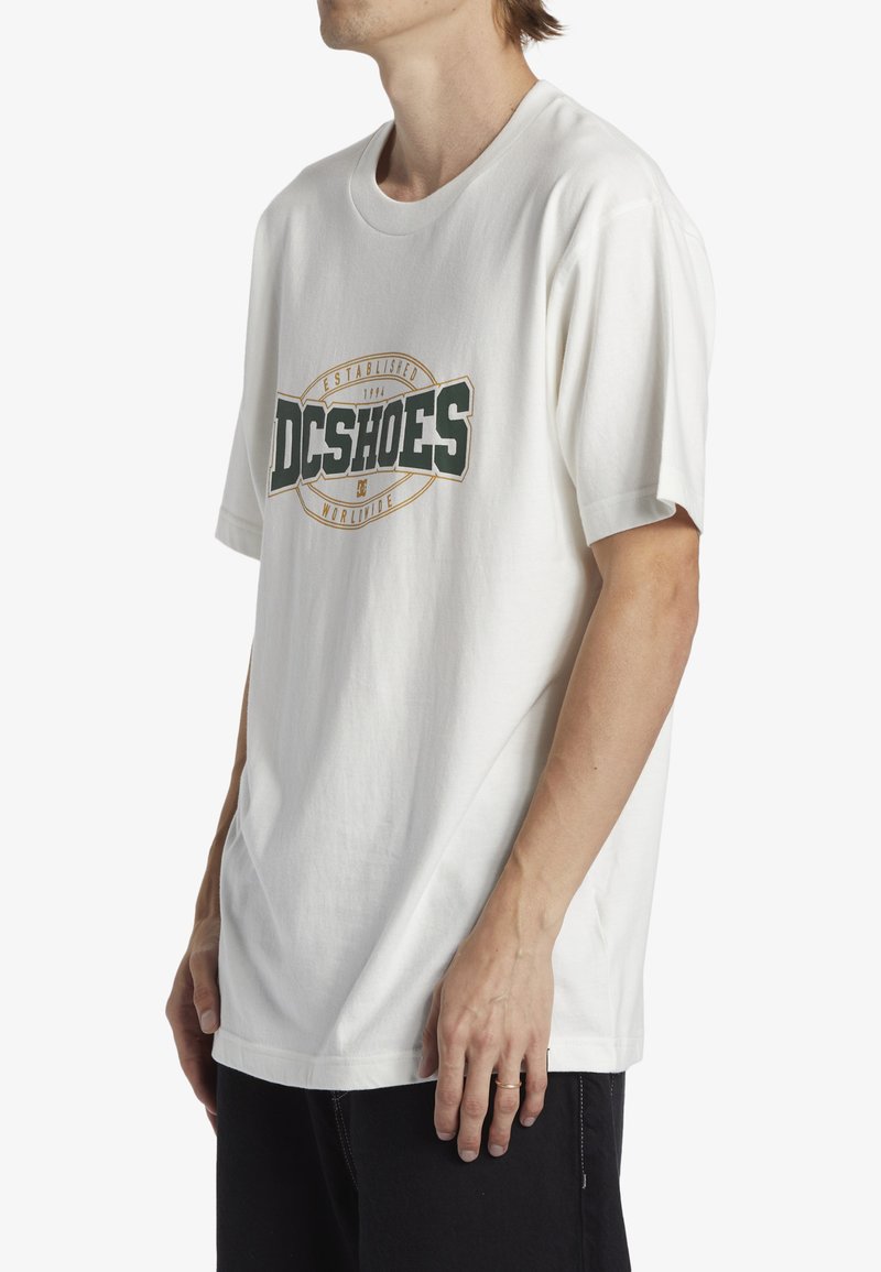 White short-sleeve t-shirt with a green and gold "DC SHOES" logo in the center. Made of cotton, featuring a relaxed fit and round neckline.