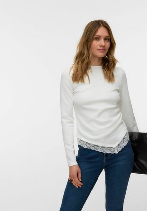 Kvinde med bølget blondt hår iført en hvid langærmet top med blonder og blå jeans, der holder en sort håndtaske.