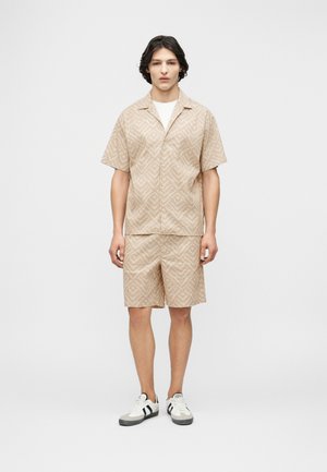 Ung mand stående, iført beige kortærmet skjorte med mønster og matchende shorts, med hvide og sorte sneakers.