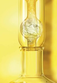 Flacon de laboratoire en verre transparent à fond rond, rempli d'un liquide jaune. Des bulles notables remontent à la surface du liquide, sur fond jaune éclatant.