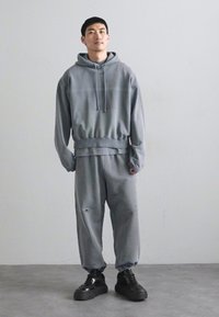 Šedá mikina s predným vreckom, skombinovaná so zodpovedajúcimi nohavicami s opotrebovanými detailmi. Čierne tenisky dotvárajú celý outfit.