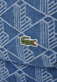 Denim stof met een geometrisch patroon in blauw en wit, met een geborduurd groen krokodil logo met een rode mond.