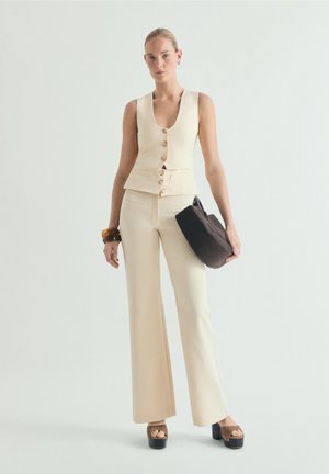 Mujer con chaleco sin mangas color crema con botones y pantalones anchos a juego, sosteniendo un bolso negro texturizado y usando sandalias de plataforma.