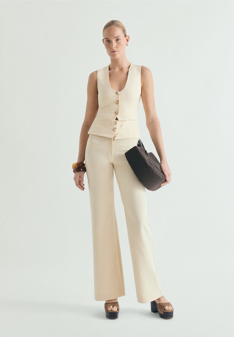 Mujer con chaleco sin mangas color crema con botones y pantalones anchos a juego, sosteniendo un bolso negro texturizado y usando sandalias de plataforma.