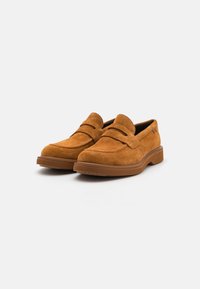 Zapatos de ante sin cordones en marrón con una textura suave, que presentan una punta redondeada y una sutil tira decorativa en el empeine.