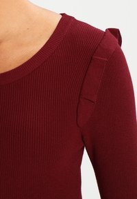 Maglione bordeaux a coste con scollatura rotonda, caratterizzato da sottili dettagli a volant sulle spalle. Tessuto testurizzato, design aderente.