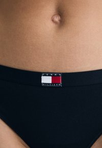 Bas de bikini noir avec une étiquette rectangulaire de logo Tommy Hilfiger, présentant des accents rouges, blancs et bleus sur la taille. Tissu lisse et extensible.