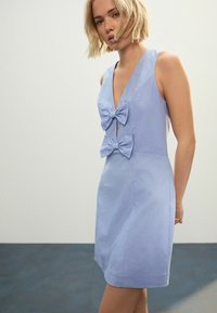 Hellblaues ärmelloses Kleid mit tiefem V-Ausschnitt und zwei großen Schleifen an der Vorderseite, aus glattem Stoff mit taillierter Passform.