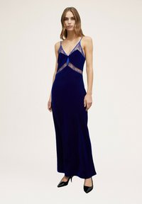 Largo vestido de terciopelo azul con acentos de encaje, escote en V profundo, tirantes ajustables y una falda fluida. Combinado con zapatos de tacón alto negros.