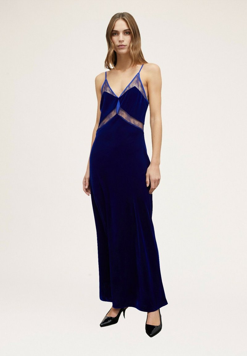 Largo vestido de terciopelo azul con acentos de encaje, escote en V profundo, tirantes ajustables y una falda fluida. Combinado con zapatos de tacón alto negros.