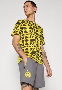 Gul- og sortmønstret kortærmet skjorte med rund halsudskæring, parret med grå shorts med et logo og sidelommer med lynlås.