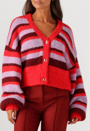 Vrouw in een fluffy, cropped cardigan met rode, paarse en donkerrode horizontale strepen en grote knopen, gecombineerd met rode broek.