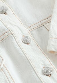 Bijeli denimski materijal s srebrnim gumbima, kontrastnim šavovima i teksturiranom površinom. Kratki kadar prikazuje detalje i završnu obradu tkanine.
