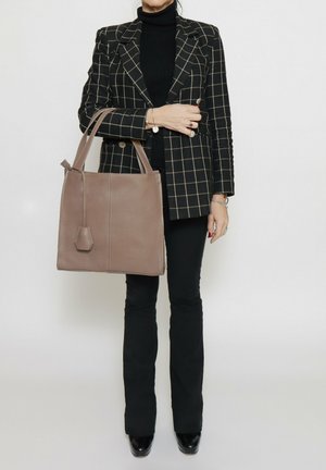 Bruine leren totebag met twee stevige handvatten, rechthoekige vorm en een gestructureerde afwerking. Draagt door een persoon in een geruit blazer.