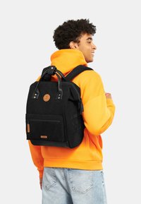 Zaino in corduroy nero con maniglia superiore e spallacci regolabili. Caratterizzato da una tasca anteriore con zip e un emblema del marchio sul davanti. Design resistente.