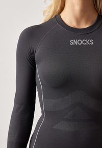 Black long-sleeve athletic top with a snug fit. Features subtle texture variations, white stitching, and branding in the upper chest area.