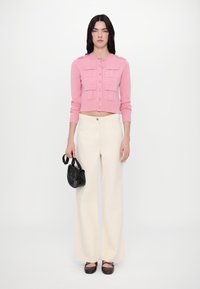 Cardigan court rose avec quatre poches avant et fermeture à boutons, associé à un pantalon large crème et un petit sac à main noir. Des chaussures marron complètent le look.