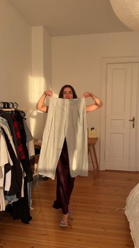 Helle graue, hochtaillierte Hose mit Falten und seitlichen Taschen. Der Stoff wirkt strukturiert. Der Hintergrund zeigt einen Kleiderständer.