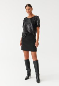 Top corto a maniche corte in paillettes nere abbinato a una gonna nera aderente. Modella che indossa stivali neri con tacco al ginocchio. Design semplice e raffinato.