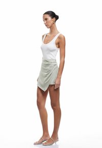 Vrouw in profiel staande, gekleed in een witte tanktop, een lichtgroene asymmetrische minirok, gouden oorringen en witte sandalen met bandjes.