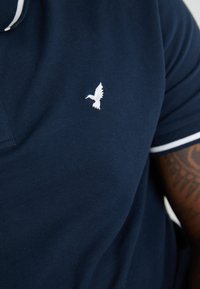 Tumesinine polo särk, millel on valged aktsendid ja vasakul rinnal tikandiga valge linnu logo. Materjalil on tekstureeritud viimistlus.