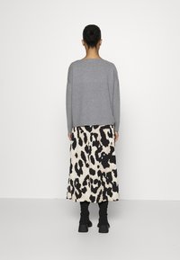 Nainen, jolla on hiukset nutturalla, pukeutunut harmaaseen neuleeseen, musta-beige leopardikuvioinen midi-hame ja mustat saappaat, seisomassa valkoista seinää vasten.