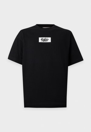 Jeune homme debout portant un t-shirt noir avec un texte graphique, un jean large bleu clair et des chaussures marron sur un fond blanc uni.