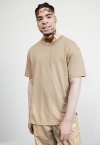 Bež majica iz 100% bombaža s okruglim izrezom i kratkim rukavima. Sadrži mali, izvezeni Nike logo u središtu. Nosena s usklađenim cargo hlačama.