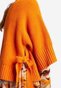 Pull en tricot orange avec manches côtelées, présentant des fentes latérales et des accents noués. Superposé sur un tissu à motifs aux nuances d'orange.
