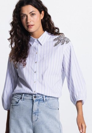 Vrouw met krullend donker haar, gekleed in een lichtblauw gestreept overhemd met knopen en zilveren bladborduursel op de schouder, en een lichtblauwe spijkerbroek.