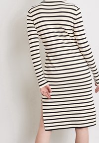 Robe tricotée rayée en crème et noir, avec des manches longues, des fentes latérales et une silhouette ajustée, mettant en valeur le motif contrastant.
