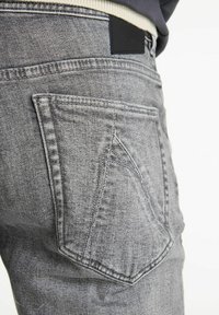 Jeans de denim gris con un bolsillo trasero con una abertura angulada distintiva, un acabado texturizado y una etiqueta de cuero negro en la cinturilla.