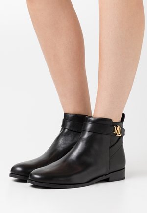 Bottes basses en cuir noir avec une sangle et un détail logo doré "RL", portées sur des jambes nues sur un fond blanc.