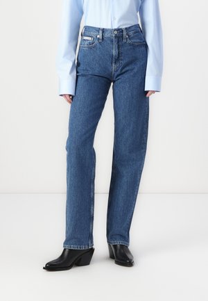 Personne portant un jean bleu droit, une chemise bleu clair à manches longues et des bottines noires à talons, debout sur un sol blanc.