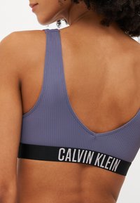 Sportovní podprsenka Calvin Klein v jemně fialové barvě, s žebrovanou texturou, černým elastickým páskem se bílým logem. Design obsahuje hluboký vykrojený zadní díl.