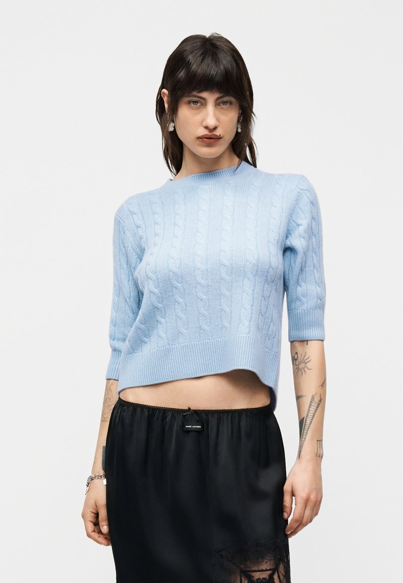 Personne portant un pull court bleu clair en maille torsadée et une jupe noire, avec des bras tatoués visibles sur un fond blanc.