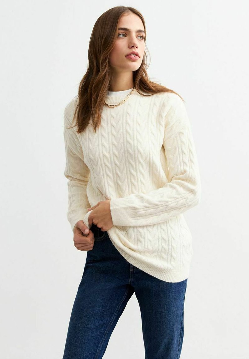 Maglione crema a lavorazione a treccia con scollo alto e polsini a coste, abbinato con jeans blu scuro. Presenta motivi testurizzati in tutto il capo.