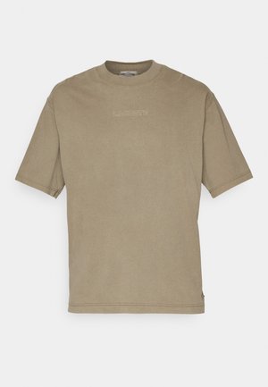 Mężczyzna ubrany w oliwkowo-zielony t-shirt z krótkim rękawem, niebieskie jeansy z podwiniętymi nogawkami i białe trampki stoi obok beżowego drewnianego krzesła z poduszką.