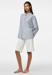 Prugasta košulja s dugim rukavima u plavoj i bijeloj boji, s ovratnikom i prednjim džepom, u kombinaciji s bijelim šortsevima do koljena i bež japankama.