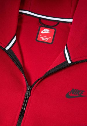 Rød Nike hoodie med sort lynlås og accenter. Har et mærke med vaskeanvisninger og et logo på brystet. Blød stofstruktur.