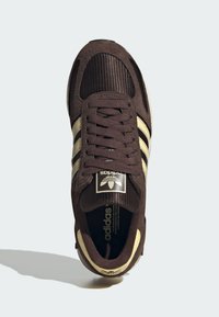 Καφέ σαουέτ αθλητικό παπούτσι με μπεζ ρίγες, ραβδωτή υφή στη μύτη και μαύρη ετικέτα με λογότυπο στη γλώσσα.