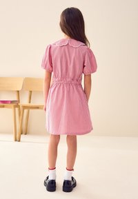 Vestito rosso a quadretti con maniche a sbuffo, collo alla Peter Pan e vita elasticizzata. Indossato con calzini bianchi e scarpe nere in vernice.